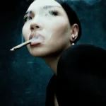 A noir-inspired close-up portrait of a woman partially obscured by cigarette smoke, lit with soft low-key lighting against a dark background. Her expression appears distant and emotionally restrained, while the grainy texture and atmospheric haze create a cinematic feeling of urban melancholy and psychological tension, by Photographer Alexey Velikorodny