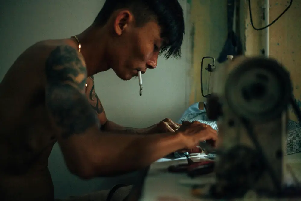 A local Southeast Asian artist, shirtless with a tattoo on his shoulder, working on handcrafts in a scene identical to a laborer processing goods at home.
